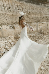 Braut steht mit fließendem ivory Boho Brautkleid ohne Spitze mit Ärmelstulpen in felsiger Natur. Kleid erhältlich bei Halona Brautmode Bodensee und Schweiz